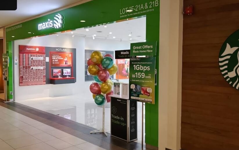 Sebuah gambar pandangan hadapan kedai runcit Maxis yang terletak di dalam pusat membeli-belah. Kedai ini mempunyai papan tanda hijau yang cerah dengan logo Maxis. Di hadapan kedai, terdapat dirian paparan yang mempromosikan 'Maxis Home Fibre' dengan harga RM159 sebulan untuk kelajuan 1Gbps