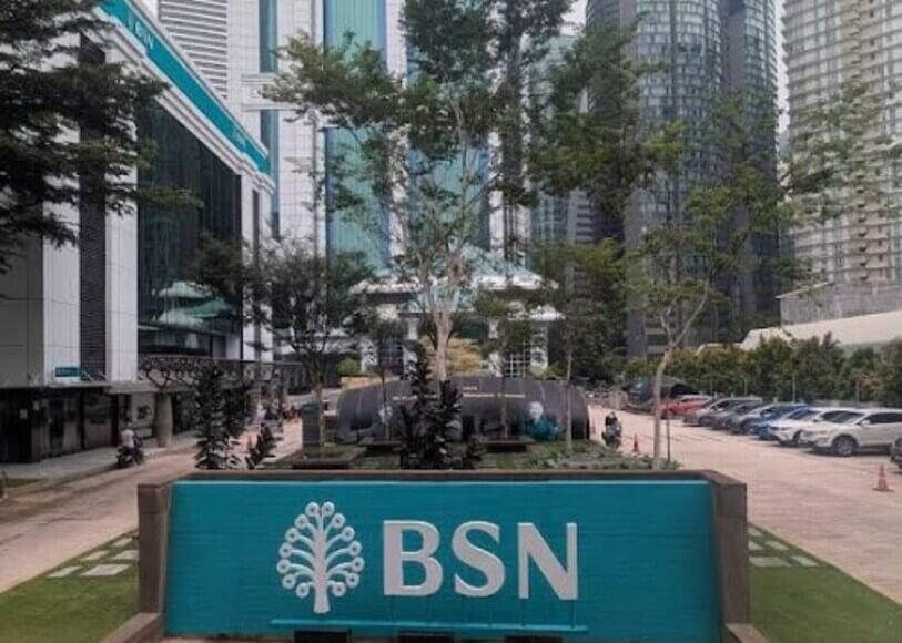 Pandangan hadapan papan tanda BSN berwarna biru turquoise di kawasan luar cawangan. Di bahagian belakang kelihatan bangunan utama BSN, kawasan tempat letak kereta, pokok-pokok, dan gedung pencakar langit
