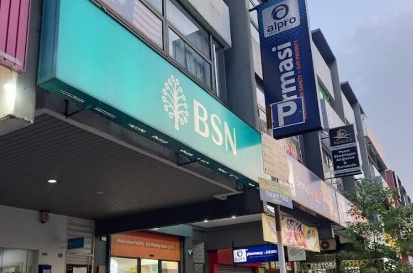 Pandangan bawah dari sudut tepi menunjukkan papan tanda hijau Bank Simpanan Nasional (BSN) dengan logo pohon, bersebelahan papan tanda gantung Farmasi Alpro di sebuah deretan bangunan kedai