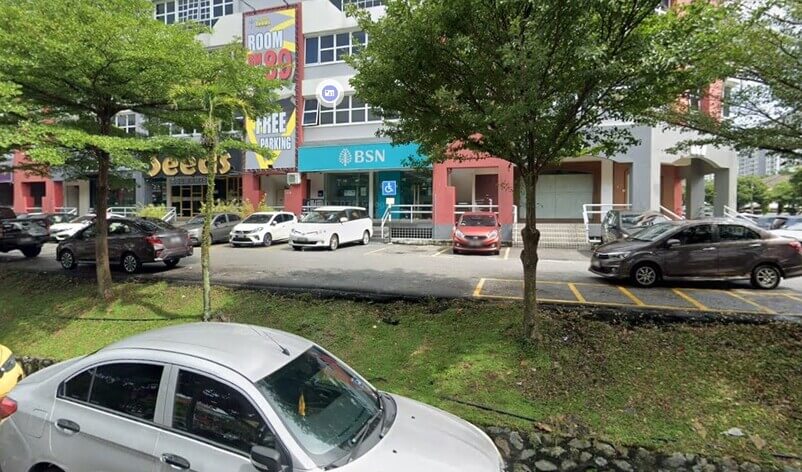 Pandangan hadapan cawangan Bank Simpanan Nasional (BSN) dengan papan tanda berwarna biru muda di tingkat bawah bangunan komersial, diapit oleh papan iklan "ROOM RM99" dan perniagaan "SEEDS", dengan barisan kereta diletakkan di hadapannya serta pokok-pokok hijau di bahagian hadapan laluan