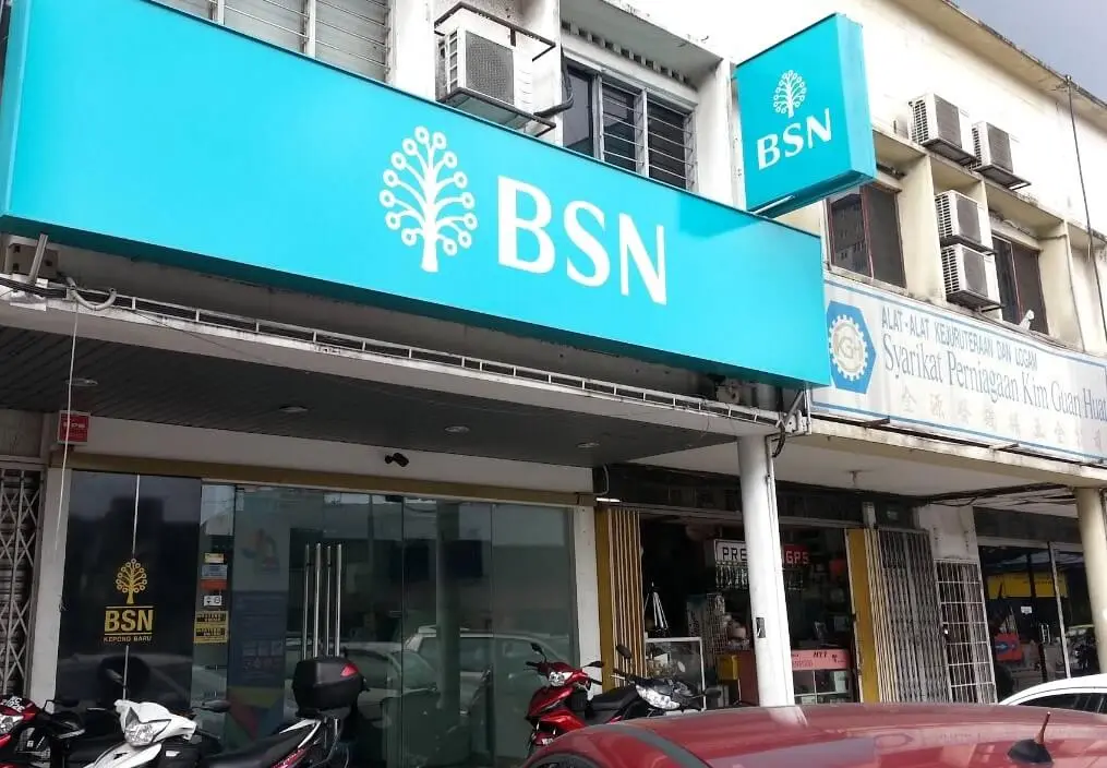Pandangan jarak dekat papan tanda utama dan papan tanda gantung BSN berwarna hijau pirus di bahagian hadapan premis cawangan BSN Kepong Baru, bersebelahan kedai Syarikat Perniagaan Kim Guan Huat