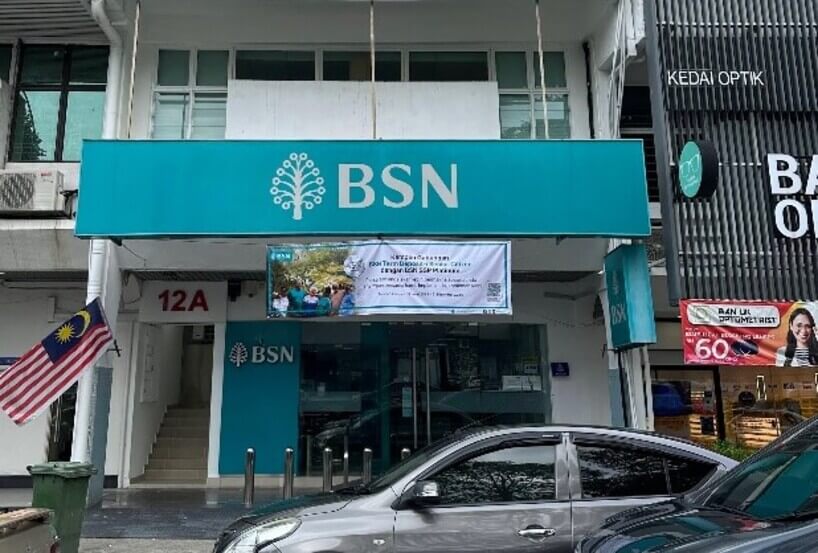 Pemandangan hadapan cawangan Bank Simpanan Nasional (BSN) dengan papan tanda berwarna biru pirus, bendera Malaysia di bahagian kiri, dan sebuah kereta di bahagian hadapan