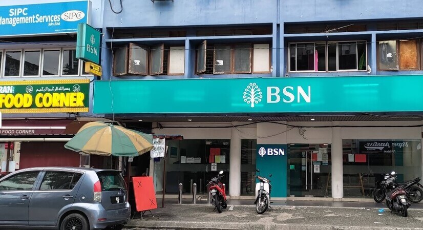Fasad kedai cawangan BSN dengan papan tanda panjang berwarna biru pirus bertuliskan logo dan nama BSN. Bangunan dua tingkat ini terletak bersebelahan restoran "Food Corner" di sebelah kiri, dengan pintu masuk kaca, papan iklan kecil, payung penjaja, serta beberapa buah motosikal dan kereta diletakkan di luar premis
