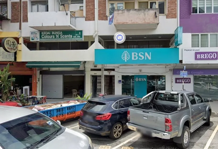 Pandangan luar cawangan Bank Simpanan Nasional (BSN) bertanda biru di deretan bangunan kedai, bersebelahan Kedai Bunga Colours 'N' Scents. Kelihatan kenderaan seperti trak pikap dan SUV diletakkan di petak tempat letak kereta di hadapan premis