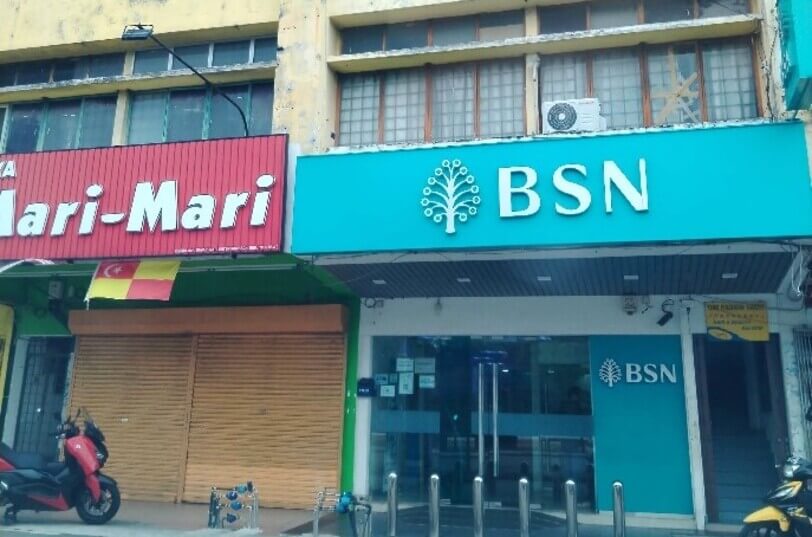 Pandangan hadapan cawangan Bank Simpanan Nasional (BSN) bertanda papan tanda biru muda, bersebelahan dengan kedai Mari-Mari yang mempunyai bendera Selangor di hadapannya