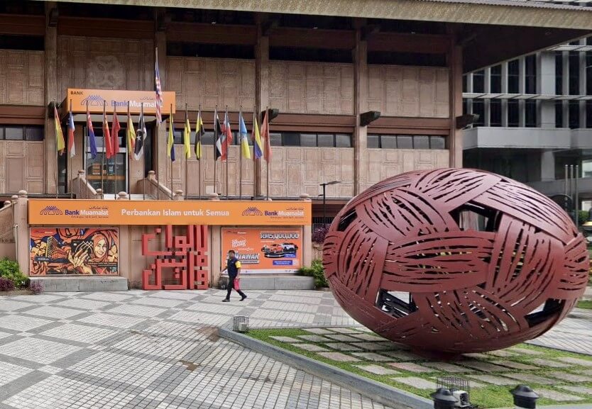 Eksterior Bank Muamalat Malaysia Berhad, menampilkan arca logam bulat besar berwarna coklat kemerahan dengan tekstur tenunan terbuka yang menyerupai bola rotan atau bola sepak takraw di latar depan. Pintu masuk bank di latar belakang mempunyai fasad oren memaparkan logo dan slogan 'Perbankan Islam untuk Semua', diapit dengan deretan bendera negara dan negeri