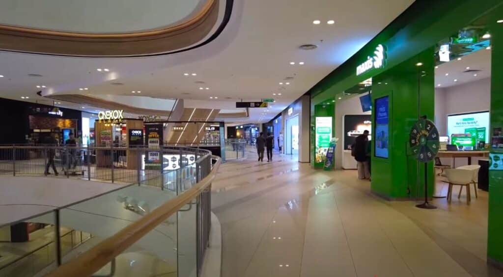 Pemandangan dalaman tingkat atas sebuah pusat beli-belah moden yang terang benderang, menunjukkan kedai Maxis Centre di sebelah kanan dengan fasad hijau terangnya