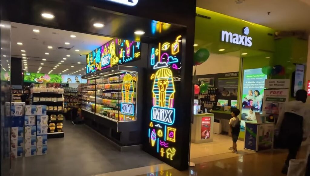 Etalase dua perniagaan bersebelahan di Sunway Pyramid. Di sebelah kiri, terdapat kedai serbaneka, lampu neon yang besar dan berwarna-warni di tiang hadapan. Di sebelah kanan, terdapat Maxis Centre, ditandai dengan logo Maxis hijau yang menarik dan penjenamaan di dinding