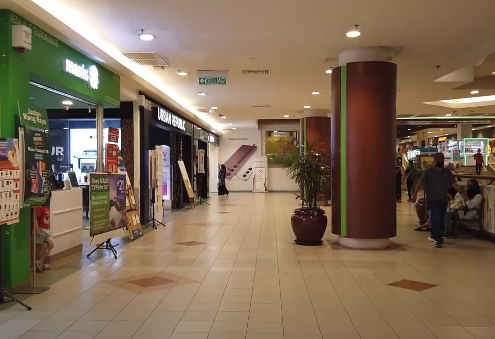 Seseorang berjalan di lorong mall yang terang benderang. Di sebelah kiri ialah kedai Maxis Centre dengan logo hijau dan paparan promosinya. Di sebelahnya ialah kotak paparan gelap kedai Urban Republic. Beberapa tiang berdiri di tengah-tengah lorong, dan di latar belakang, orang duduk di bangku