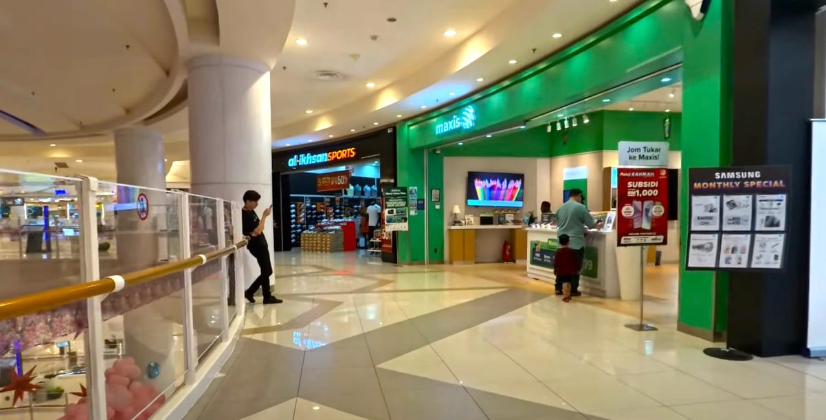Kedai Maxis Centre dengan fasad hijau terang di dalam pusat beli-belah, Aeon Shah Alam. Kedai itu mempunyai meja dan pameran berwarna putih, dengan kakitangan dan pelanggan berhampiran kaunter