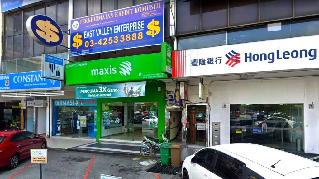 Foto kedai komersil. Bahagian tengah hadapan mempunyai pusat khidmat mudah alih Maxis berwarna hijau terang dengan papan tanda besar bertulis "PERCUMA 3X GANDA Kelajuan Internet". Di atas papan tanda Maxis terdapat papan tanda besar berwarna biru dan kuning yang bertulis East Valley Enterprise, dengan perkataan "PERKHIDMATAN KREDIT KOMUNITI"
