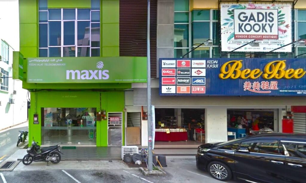 Kompleks membeli-belah komersial dua tingkat. Di sebelah kiri, Maxis Centre dicat dengan warna hijau terang khas Maxis. Tingkat bawah mempunyai kotak paparan kaca, dan tingkat atas mempunyai tingkap dengan logo dan teks Maxis. Di sebelah kanan Maxis Centre terdapat dua lagi tingkap kedai
