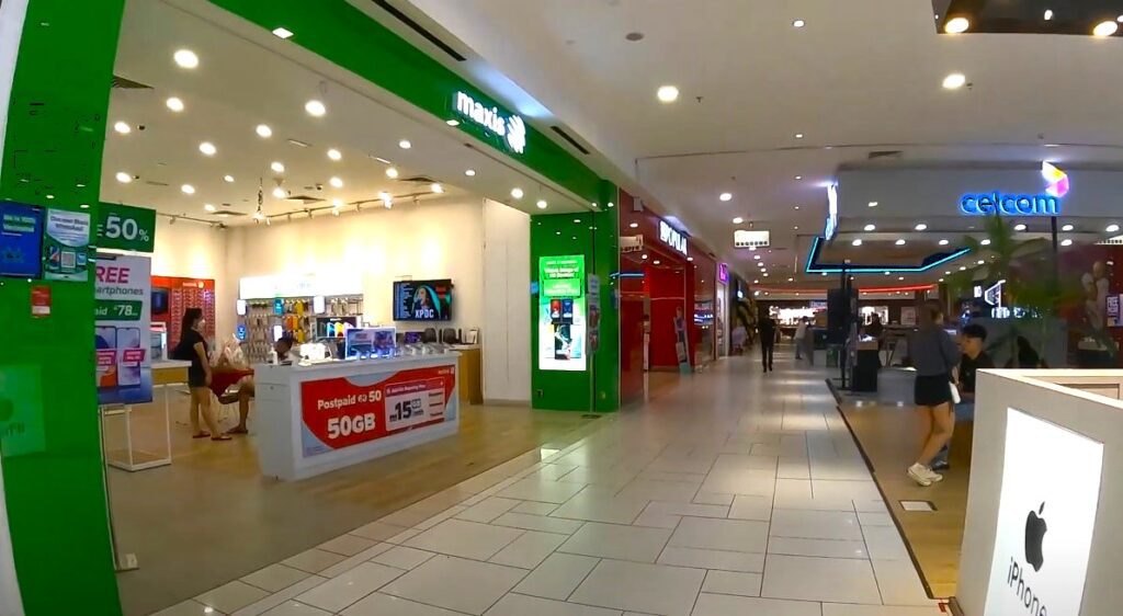Bahagian dalam lorong pusat membeli-belah yang diterangi terang. Di sebelah kiri ialah kedai pembekal perkhidmatan mudah alih Maxis dengan papan tanda hijau dan kaunter putih yang memaparkan telefon pintar. Terdapat beberapa orang di dalam kedai dan papan tanda bertulis ""Postpaid 5G 50GB" dengan harga RM78