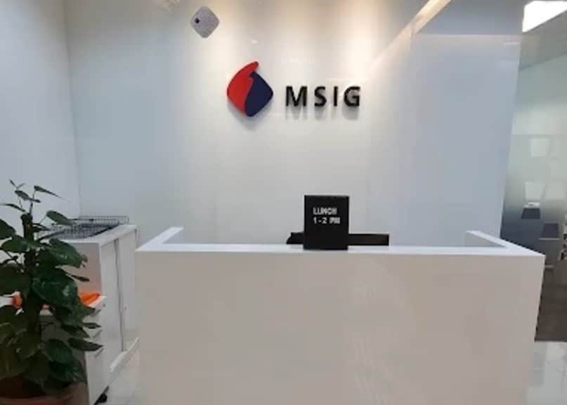 Kawasan penerimaan pejabat korporat. Fokus utama ialah meja penyambut tetamu berwarna putih, moden dan minimalis. Di atas meja, papan tanda hitam kecil bertulis "LUNCH 1-2 PM" dalam teks putih. Di belakang meja, dinding putih memaparkan logo dan nama syarikat. Logo ialah reka bentuk merah dan biru yang digayakan, diletakkan di sebelah kiri nama syarikat. Nama, "MSIG," ditulis dalam huruf besar, hitam, tiga dimensi