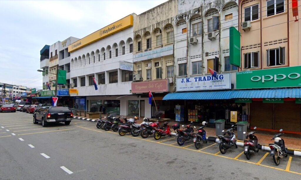 Gambar menunjukkan bahagian hadapan pejabat Maybank Mersing yang terdiri daripada gabungan beberapa kiosk. Terletak di kawasan komersial dengan banyak kiosk tiga tingkat di sekelilingnya. Bangunan pejabat itu dicat putih dan mempunyai papan nama kuning besar yang dipasang di atas