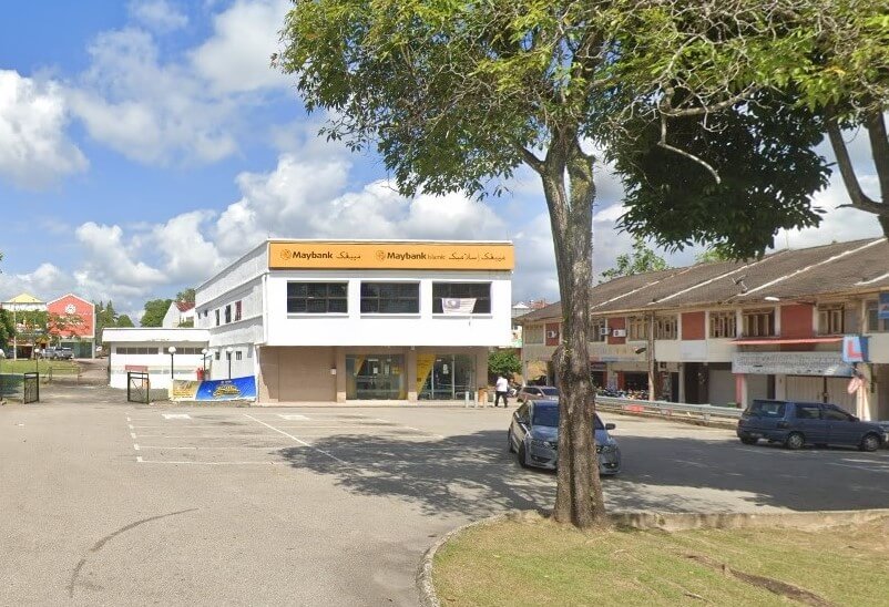 Maybank dua tingkat di Muadzam Shah, Pahang, Malaysia, dengan bumbung rata dan dicat putih dengan jalur kuning tersendiri di bahagian atas, memaparkan logo Maybank dan perkataan "Maybank" dan "Maybank Islamic." Kawasan parkir berturap berada di latar depan, dengan beberapa kereta parkir di sebelah kanan. Sebatang pokok yang besar dan rendang berdiri teguh di tengah-tengah bingkai itu