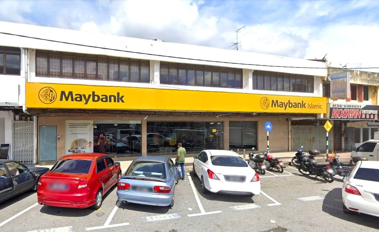 Gambar menunjukkan lokasi pejabat cawangan Maybank Skudai yang berada di kawasan komersial dengan banyak kios dua tingkat di sekelilingnya. Bangunan itu dicat putih, gabungan coklat dan kuning, dan mempunyai papan nama di hadapan