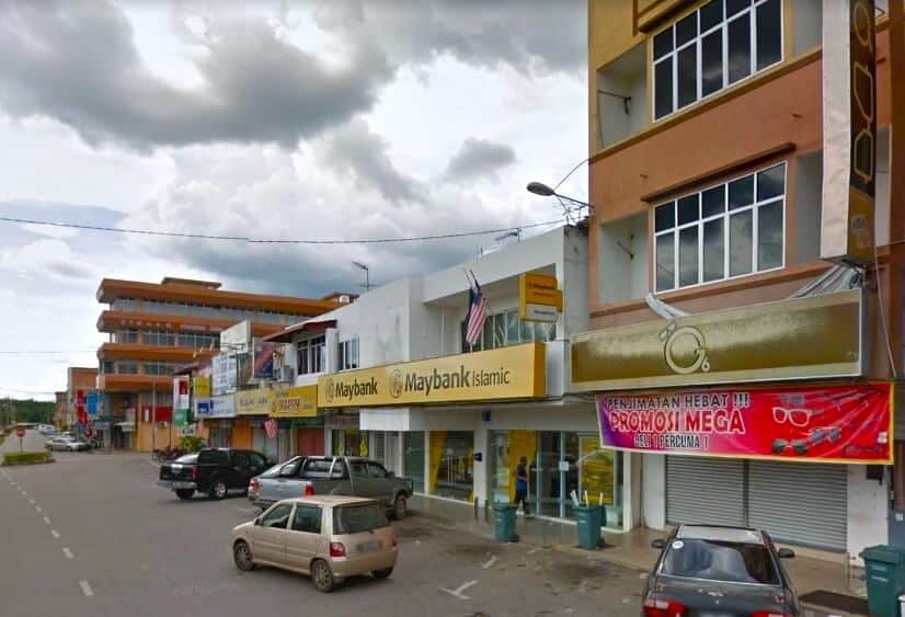 Deretan bangunan komersial kecil di Pahang, Malaysia, setinggi dua hingga tiga tingkat dan diperbuat daripada konkrit bercat, dengan pelbagai papan tanda dan sepanduk. Jalan dengan kereta yang parkir dan laluan pejalan kaki yang sempit kelihatan di latar depan. Bangunan utama adalah cawangan Maybank dengan papan tanda kuning bertulis "Maybank" dan "Maybank Islamic" berwarna hitam. Bangunan di sebelah kanan mempunyai sepanduk merah besar dengan huruf putih dan kuning serta imej cermin mata, mengiklankan promosi