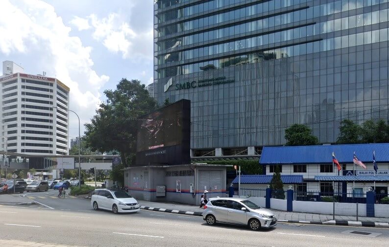 Pemandangan jalanan kawasan bandar yang sibuk menunjukkan bangunan pencakar langit kaca moden dengan perkataan "SMBC" dan "BANKING CORPORATION" pada fasadnya, mengenal pasti ia sebagai bangunan Sumitomo Mitsui Banking Corporation (SMBC) di Malaysia
