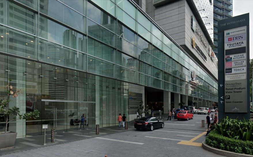 Pintu masuk ke bangunan Vista Tower moden bertingkat dengan fasad kacanya, menunjukkan akses aras jalan ke kompleks. Papan tanda di sebelah kanan menunjukkan lokasi 'The Intermark' dan beberapa perniagaan termasuk 'DBS,' 'Tsutaya Books,' 'Makan Kitchen,' 'Cellar Door,' dan 'Tosca,' serta 'DoubleTree by Hilton' di 'Retail Podium Vista Tower.'