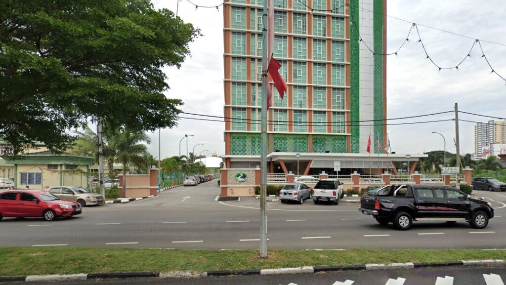 Foto luaran paras mata ini merakam pintu masuk dan fasad bawah bangunan pejabat tinggi dan moden, Kantor Air Kelantan Sdn Bhd. Bangunan berbilang tingkat itu menampilkan jeriji tingkap berwarna coklat jingga dan aksen biru menegak yang berulang, memberikan rupa jubin yang tersendiri