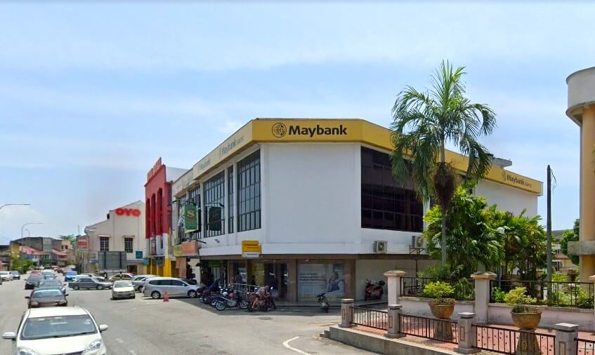 Gambar siang hari dari aras jalan menunjukkan cawangan Maybank Batu Gajah, sebuah bangunan dua tingkat di Malaysia dengan tanda kuning dan hitam yang tersendiri. Bank itu terletak di sudut lot, dikelilingi oleh bangunan komersial lain, termasuk bangunan merah dengan tanda OYO