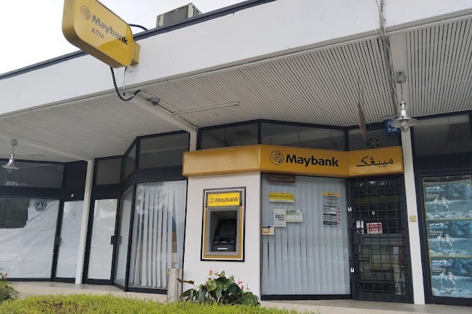 Bahagian luar cawangan kecil Maybank yang terletak di Bukit Fraser, Malaysia. Bangunan ini mempunyai fasad rata, putih pucat dengan bumbung logam beralun. Logo Maybank, yang memaparkan kepala harimau, dipaparkan pada papan tanda kuning di atas pintu masuk, dan nama "Maybank" ditulis dalam bahasa Inggeris dan Arab. ATM kelihatan di ceruk kecil di sebelah kiri pintu masuk utama