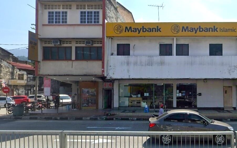 Gambar pandangan hadapan, dua bangunan komersial di jalan, dengan bangunan di sebelah kanan adalah cawangan Maybank. Bangunan Maybank dua tingkat dan dicat putih dengan papan tanda kuning yang mencolok bertulis "Maybank" dan "Maybank Islamic" dalam huruf hitam. Lantai bawah mempunyai tingkap kaca besar yang mendedahkan bahagian dalam bank