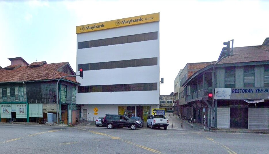 Sebuah bangunan putih empat tingkat dengan papan tanda kuning dan putih "Maybank" dan "Maybank Islamic" di atas berdiri di sudut jalan. Ia mempunyai tingkap mendatar panjang berwarna gelap pada setiap tingkat dan pintu masuk kaca di tingkat bawah. Bangunan di sebelah kiri mempunyai bumbung jubin berwarna coklat kemerahan, manakala bangunan di sebelah kanan, sebuah restoran, mempunyai fasad kayu hijau kelabu dengan balkoni