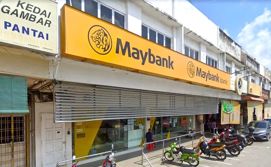 Gambar cawangan Maybank, memaparkan rumah kedai dua tingkat dengan papan tanda Maybank kuning dan hitam yang menyerlah di hadapan. Perkataan "Maybank" dan "Maybank Islamic" jelas kelihatan pada papan tanda itu. Beberapa motosikal dan sebuah kereta parkir di hadapan bangunan di jalan. Di sebelah kiri bank, terdapat papan tanda untuk "Kedai Gambar Pantai."