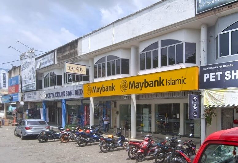 Sebuah bangunan putih dua tingkat dengan tingkap besar di tingkat atas. Di tingkat bawah, terdapat papan tanda kuning besar dengan huruf hitam "Maybank" dan "Maybank Islamic." Di bawah papan tanda adalah pintu masuk bank, dan beberapa motosikal parkir di hadapan bangunan