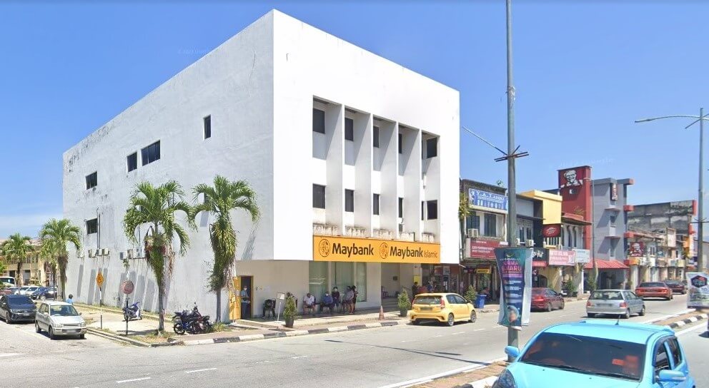 Gambar siang hari bahagian luar bangunan putih tiga tingkat dengan logo Maybank di tingkat bawah. Bangunan ini mempunyai bumbung rata dan beberapa tingkap segi empat tepat, beberapa daripadanya tersembunyi. Di hadapan bank, beberapa orang duduk di bangku, dan beberapa pokok palma berjajar di kaki lima