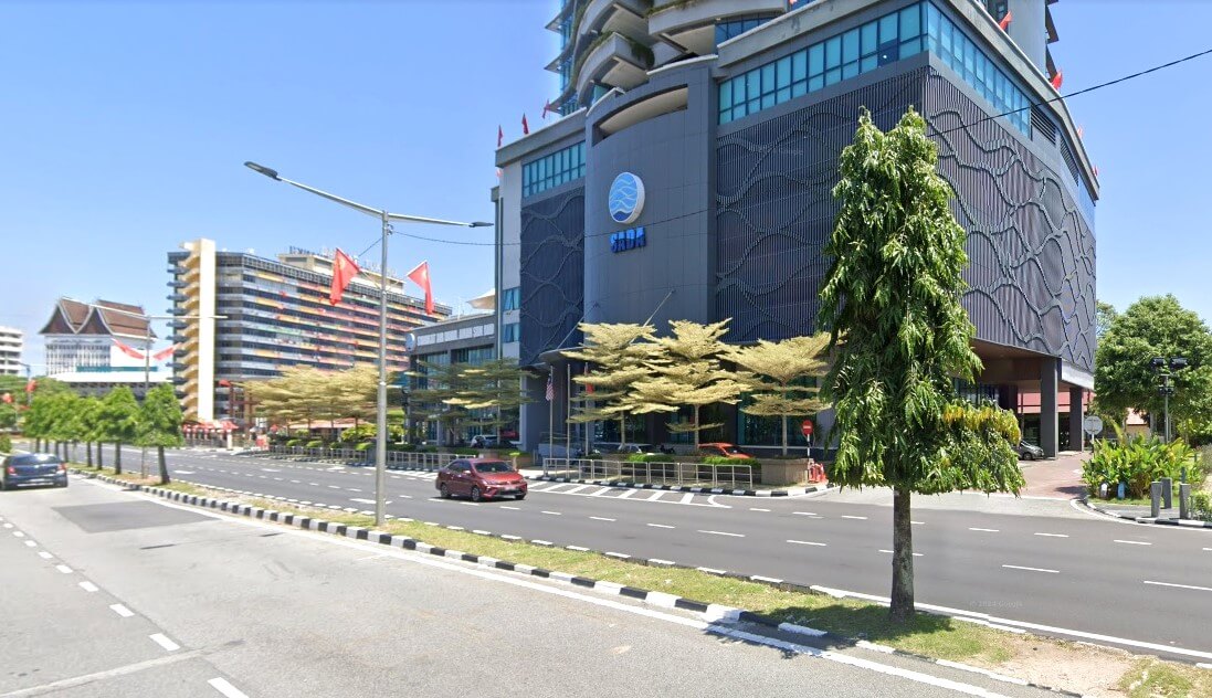 Sebuah bangunan moden bertingkat dengan fasad gelap dan tingkap pemantul, menampilkan logo biru bulat besar di hadapan, dikenal pasti sebagai Syarikat Air Darul Aman Sdn. Bhd. (SADA). Bangunan ini terletak agak jauh dari jalan berbilang lorong yang luas