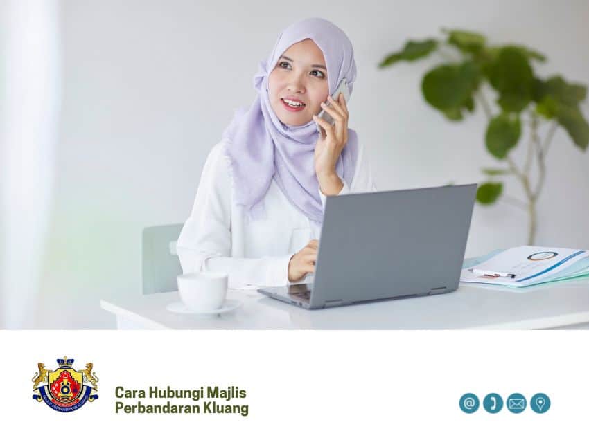 Seorang wanita memakai baju atasan putih dan tudung ungu muda sedang bercakap di telefon bimbitnya sambil mengalihkan pandangan dari komputer riba yang terbuka di atas mejanya. Imej itu juga mengandungi logo dan teks di bahagian bawah: "Cara Hubungi Majlis Perbandaran Kluang" berserta ikon kenalan (e-mel, telefon, lokasi dan tanda tanya)