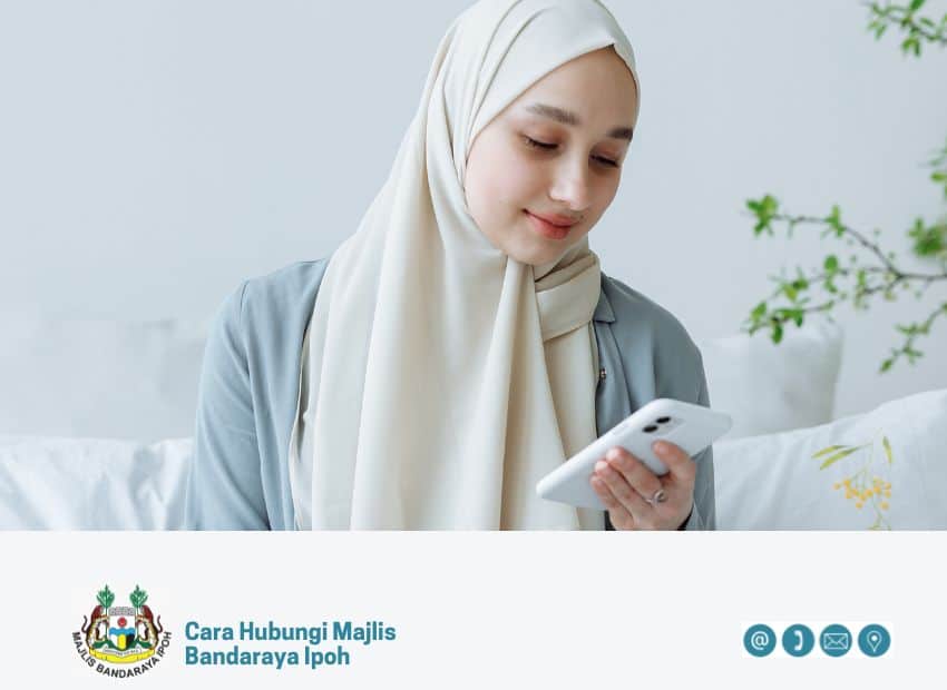 Seorang wanita muda merenung telefon pintar putihnya, memegangnya di tangannya di dalam rumah. Di bawah imej, terdapat logo Majlis Bandaraya Ipoh dan teks "Cara Hubungi Majlis Bandaraya Ipoh", berserta ikon hubungan (e-mel, telefon, lokasi)