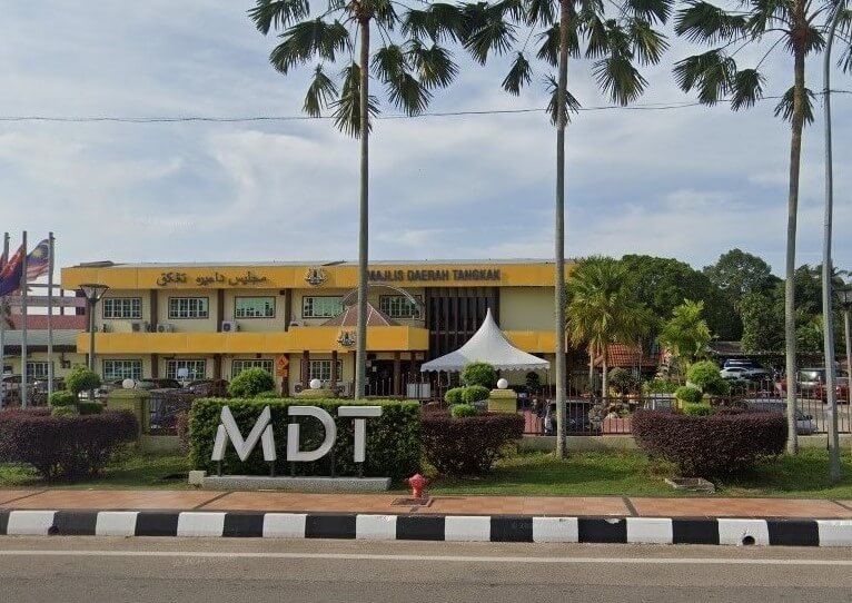 Bahagian luar bangunan Majlis Daerah Tangkak kuning dua tingkat itu memaparkan papan tanda besar "MDT" di latar depan yang tersusun rapi, dibingkai oleh pokok palma yang tinggi