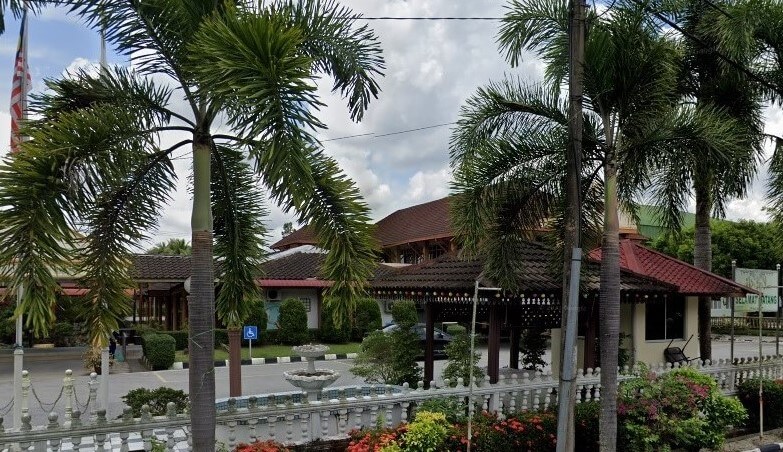 Bangunan Majlis Daerah Ketereh, yang mempunyai bumbung gaya tradisional, dikelilingi oleh dedaunan tropika yang subur, termasuk beberapa pokok palma tinggi di latar depan, dengan pagar putih hiasan dan air pancut kecil kelihatan