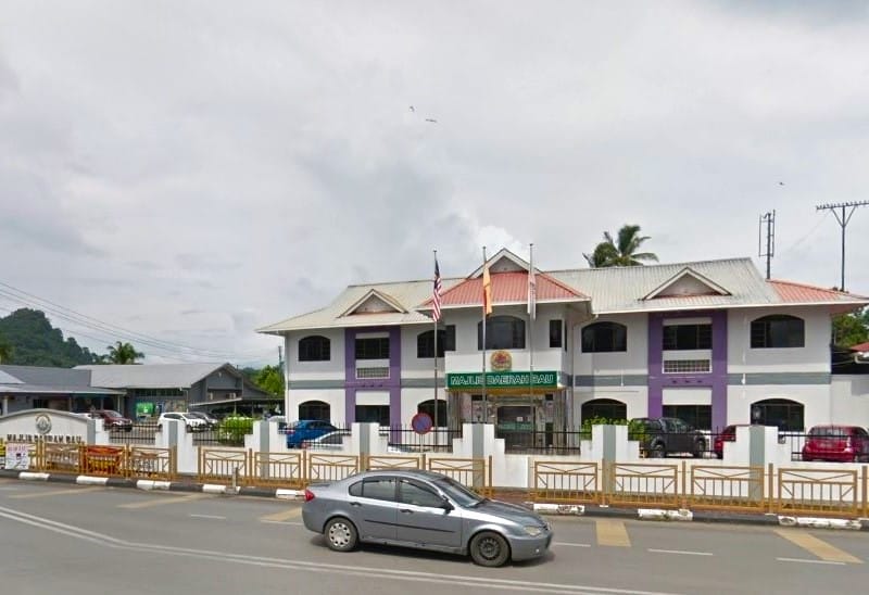 Bahagian luar bangunan Majlis Daerah Bau (MDB). Reka bentuk bangunan dua tingkat dengan cat putih dan gabungan warna ungu serta pagar yang diperbuat daripada trellis besi dengan cat kuning keemasan. Terdapat papan tanda dengan perkataan Majlis Daerah Bau dalam huruf besar di atas pintu masuk pejabat
