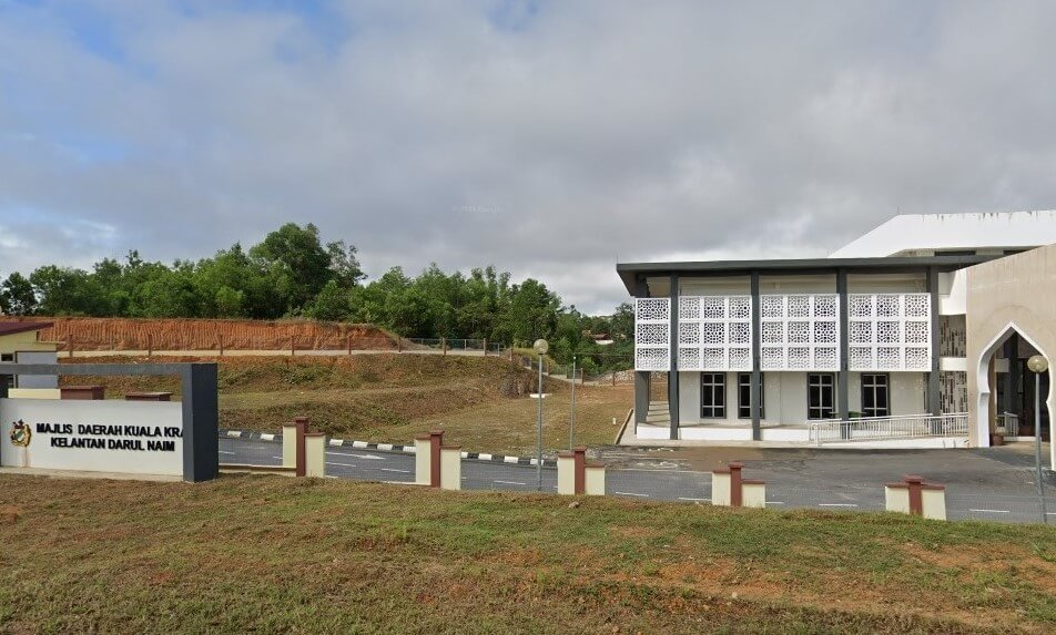 Bangunan kerajaan dua tingkat moden dengan fasad bercorak putih di sebelah kanan. Di latar depan sebelah kiri terdapat papan hitam putih bertulis "MAJLIS DAERAH KUALA KRAI KELANTAN DARUL NAIM."