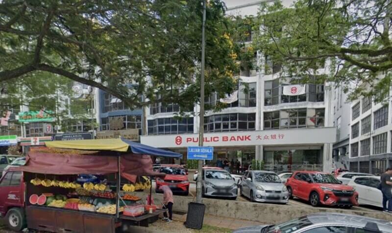 Pemandangan jalan di hadapan cawangan Public Bank Bukit Damansara dengan trak jualan buah-buahan di bahagian hadapan dan kereta yang diletakkan di tepi jalan.