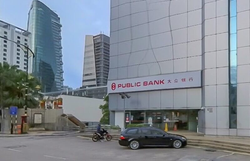 Pandangan hadapan bangunan cawangan Public Bank di Johor Bahru dengan papan tanda cawangan, sebuah kereta hitam dan penunggang motosikal di hadapannya.