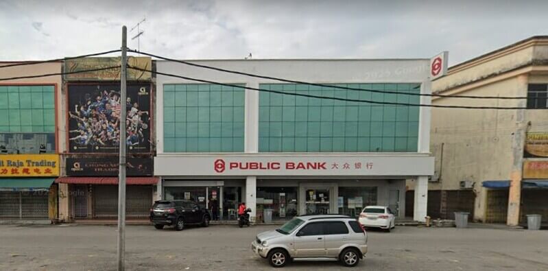 Pemandangan hadapan cawangan Public Bank di Parit Raja dengan papan tanda berwarna merah dan putih serta tulisan Cina. Bangunan dua tingkat ini mempunyai panel tingkap kaca hijau di tingkat atas, dengan beberapa buah kereta diletakkan di kawasan tempat letak kereta di hadapannya.