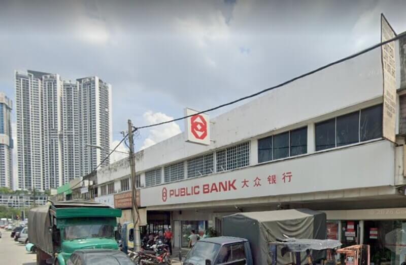 Pemandangan dari tepi jalan yang menunjukkan cawangan Public Bank Segambut di kawasan kedai dua tingkat, lengkap dengan papan tanda "PUBLIC BANK" dan tulisan Cina. Kelihatan lori dan kenderaan diletakkan di hadapan kedai, manakala bangunan kediaman tinggi bertingkat-tingkat terserlah.