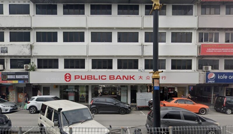 Pandangan hadapan cawangan Public Bank Ulu Tiram di sebuah bangunan kedai tiga tingkat dengan papan tanda utama serta kawasan tempat letak kereta di bahagian hadapan.