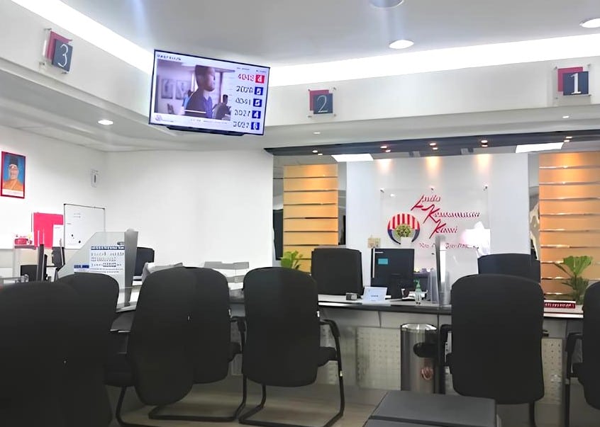 Ruang menunggu di pejabat KWSP Kulim. Monitor besar yang dipasang pada dinding memaparkan paparan digital bernombor, mungkin sistem beratur. Di bawah monitor terdapat beberapa meja dan kerusi kosong, menghadap ke arah kamera. Pejabat itu mempunyai dinding putih, dan tanda besar dengan logo boleh dilihat di belakang meja.