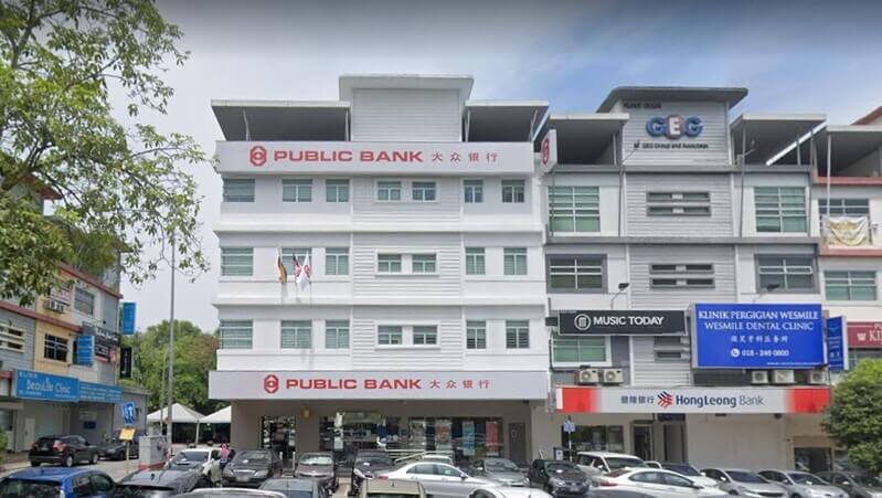 Pandangan hadapan bangunan cawangan Public Bank Bukit Jalil empat tingkat di kawasan kedai dengan papan tanda berwarna merah dan putih, berserta deretan kenderaan yang diletakkan di bahagian hadapan.