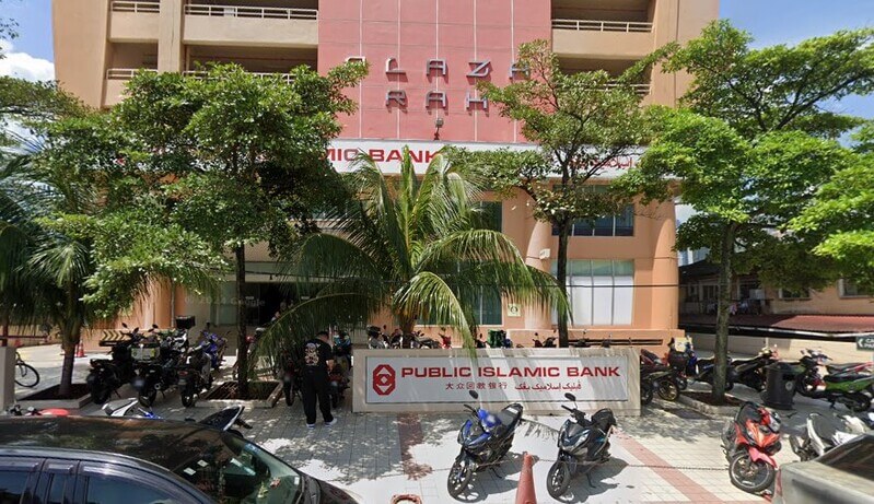 Pemandangan bahagian luar cawangan Public Bank Kampung Baru yang terletak di tingkat bawah bangunan Plaza Rah. Kelihatan tanda nama bank, papan tanda besar Plaza Rah pada dinding merah di atasnya, pokok-pokok hijau serta barisan motosikal di hadapan bangunan.