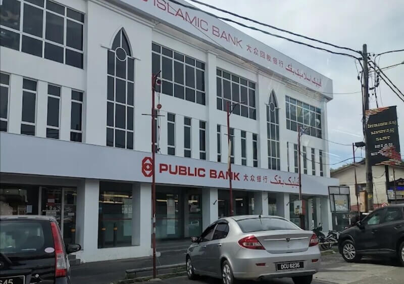 Bangunan cawangan Public Bank Kota Bharudan Public Islamic Bank berwarna putih dengan papan tanda merah dan tulisan Jawi, serta sebuah kereta Proton Persona perak diletakkan di hadapannya.