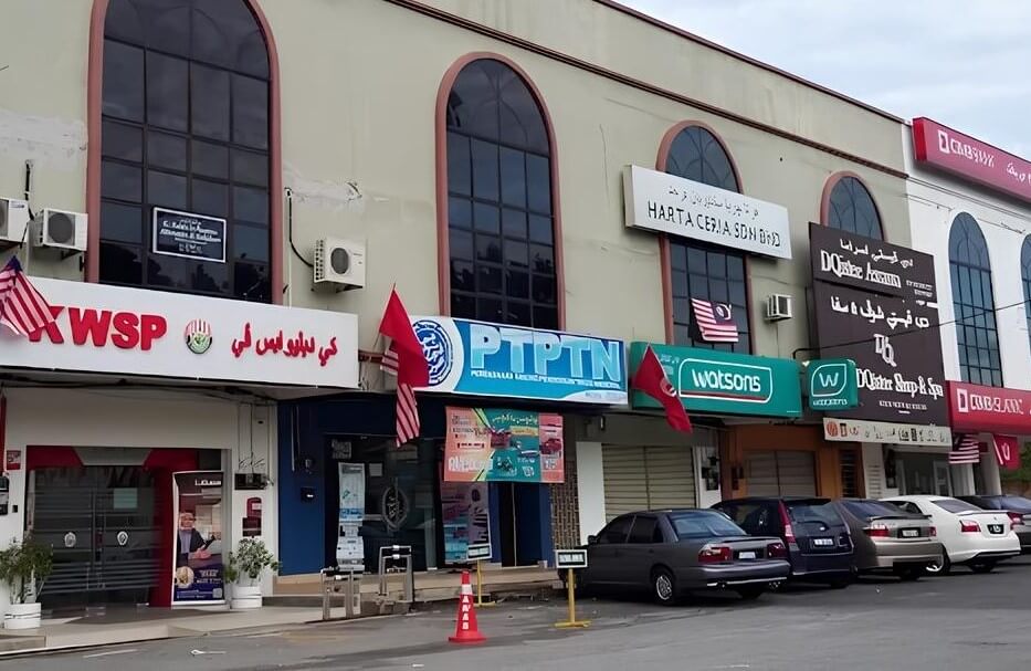 Deretan muka kedai yang saling berkait. Bangunannya berwarna krim dengan tingkap melengkung di tingkat dua. Beberapa bendera Malaysia dipamerkan di hadapan kedai. Dari kiri ke kanan, kedai-kedai tersebut ialah: KWSP (papan tanda merah putih dengan logo KWSP), PTPTN, Watsons (papan tanda hijau untuk peruncit kesihatan dan kecantikan Watsons)