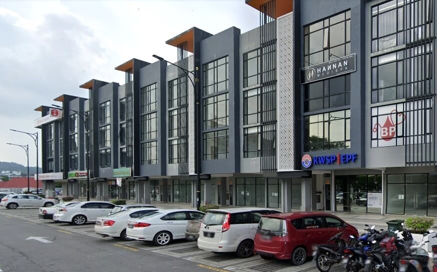 Deretan bangunan komersial berbilang tingkat moden dengan tingkap besar dan fasad kelabu-hitam. Di latar depan, kereta parkir di atas jalan asfalt yang bercerun. Di latar belakang, kelihatan bukit hijau dan langit mendung. Papan tanda KWSP EPF jelas kelihatan pada salah satu bangunan